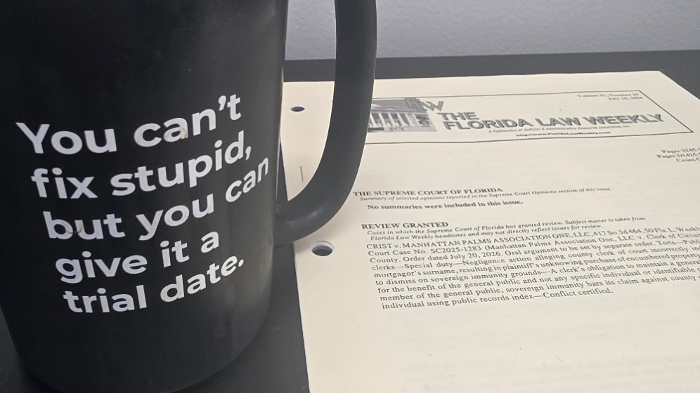 A black coffee mug reading "You can't fix stupid, but you can give it a trial date" on a desk beside an open issue of The Florida Law Weekly